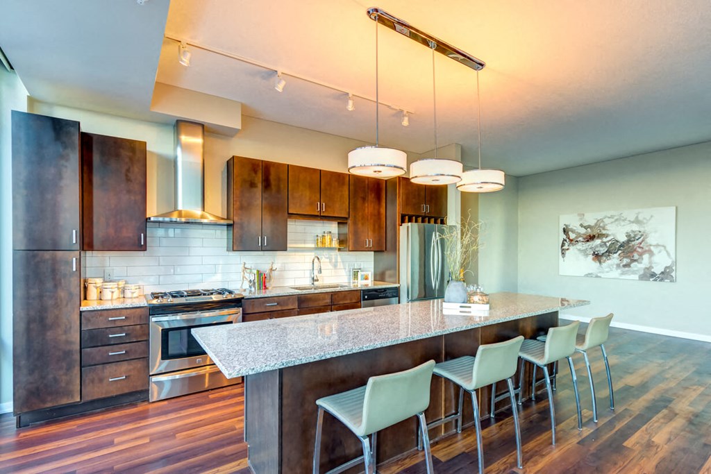Contemporary kitchen here at Nic on 5th Apartment Homes with dark wood cabinets, stainless steel appliances, and a long granite island. Pendant lights and abstract art add elegance.