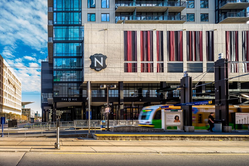 Modern urban scene with a glass-fronted building, "The Nic on 5th Apartment Homes," featuring red accents. A light rail train passes in front under a blue sky.