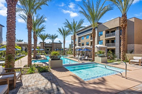 Outdoor area here at Reflect at Dobson Ranch Apartments with palm trees and a sparkling pool under a clear blue sky. Lounge chairs and umbrellas create a relaxing, tropical atmosphere.