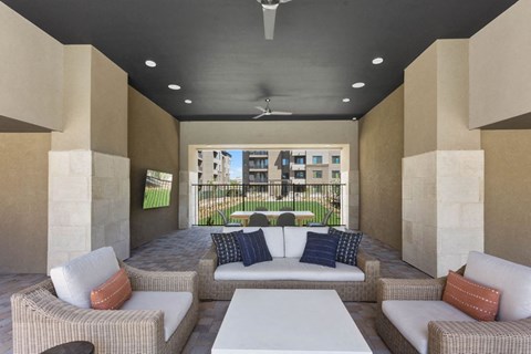 Outdoor patio area here at Reflect at Dobson Ranch Apartments with wicker furniture, colorful cushions, and a central white table. Background shows modern buildings and a grassy courtyard.