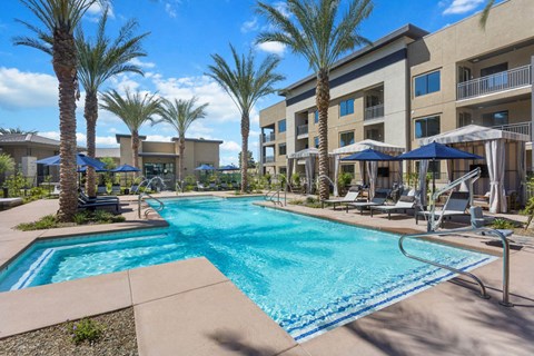 Luxurious pool area here at Reflect at Dobson Ranch Apartments with palm trees, cabanas, and lounge chairs under blue umbrellas. Modern apartment building in the background under a sunny sky.