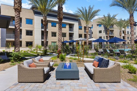 Outdoor seating area here at Reflect at Dobson Ranch Apartments with wicker sofas and colorful cushions surrounds a modern fire pit. Tall palm trees and a pool with umbrellas are in the background.