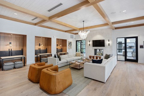 Modern lobby here at Reflect at Dobson Ranch Apartments with white walls, wood ceiling beams, and light wood flooring. White sofas, brown chairs, and a central coffee table create a cozy, elegant space. A TV and fireplace are set against a textured wall, while booths with striped cushions are nearby. Large windows allow natural light.