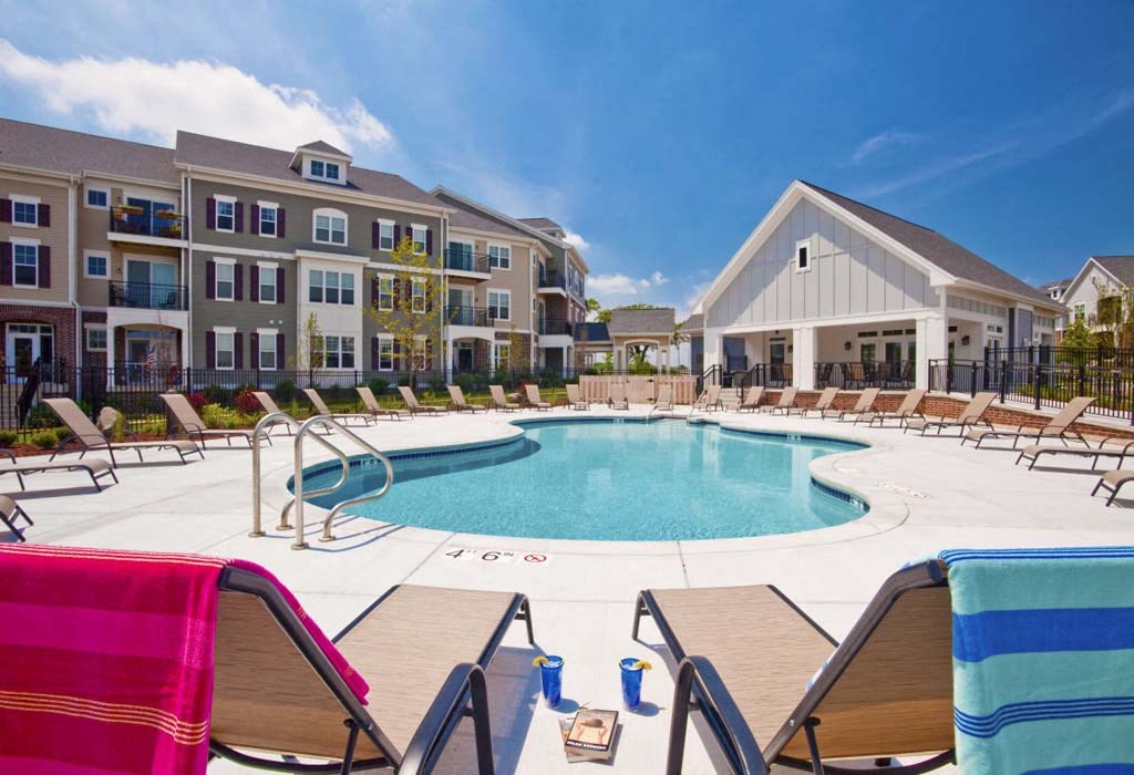 Modern Glacier Valley apartment complex with a central swimming pool, surrounded by lounge chairs and two colorful towels. Bright, sunny day with clear blue skies.
