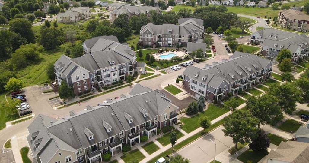Aerial view of Glacier Valley Apartments with multiple rows of modern apartment buildings, manicured lawns, and a central swimming pool. Lush green trees surround the area, creating a serene and organized atmosphere.