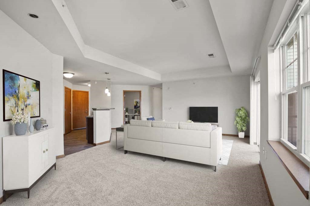 Spacious living room with neutral tones, featuring a large white sofa, a TV, and a plant in the corner. Sunlight streams through wide windows, creating a calm atmosphere. Glacier Valley at Reston Heights.