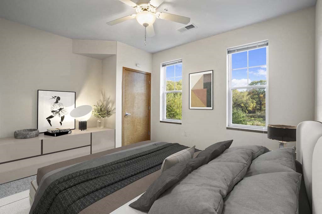 Modern bedroom at Glacier Valley Apartments with a ceiling fan, light gray walls, and two tall windows showing a green landscape. A cozy bed with gray bedding is in the foreground. A minimalist sideboard holds a sculpture, plant, and abstract art, creating a calm, serene atmosphere.