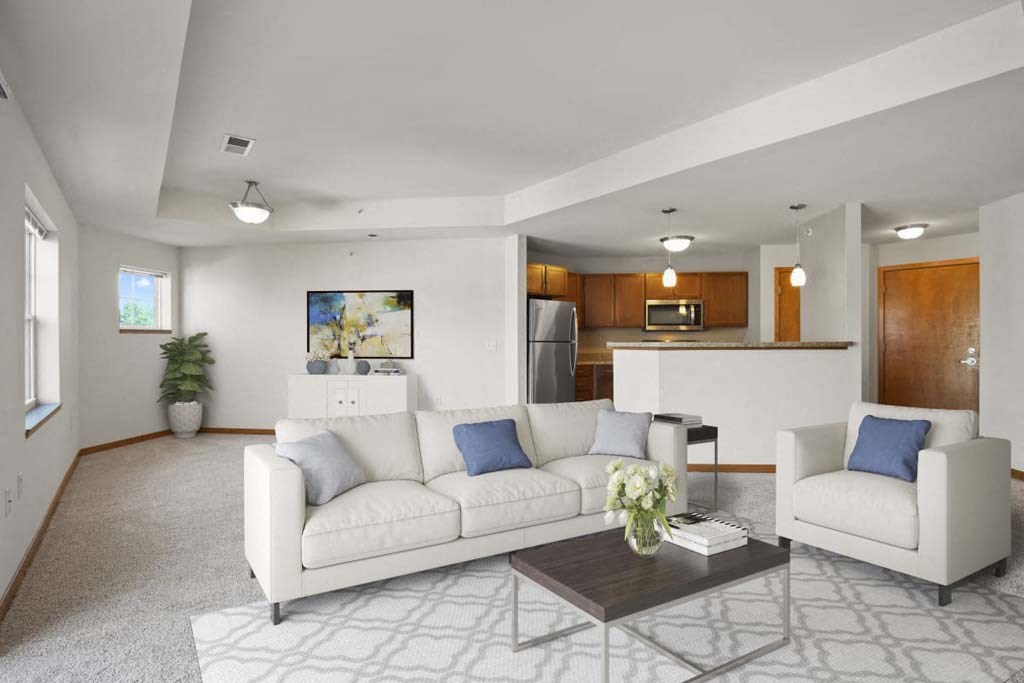 Modern living room with white sofas and gray pillows, a dark wooden coffee table, and a patterned rug. Open kitchen with wood cabinets and stainless steel appliances in the background. Bright and inviting atmosphere. Glacier Valley at Reston Heights Apartments.