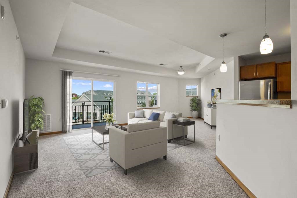 Spacious modern living room at Glacier Valley at Reston Heights Apartments with neutral tones, featuring a plush white sofa, glass coffee table, large windows, balcony view, and a sleek kitchen area.