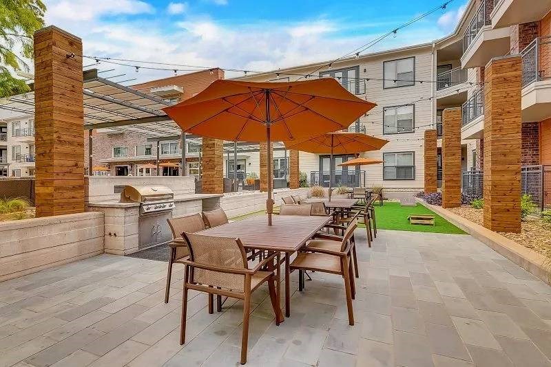 Modern courtyard here at Skyline at Farmers Market Apartment Homes with large orange umbrellas over wooden dining tables, surrounded by brick columns. A grill is in the background, evoking a relaxed, inviting atmosphere.