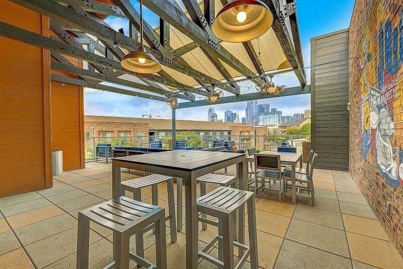 Modern rooftop patio here at Skyline at Farmers Market Apartment Homes with wooden tables, high stools, and industrial lighting. Overlooking a cityscape, the area is vibrant and inviting for social gatherings.