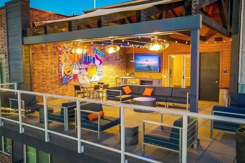 Outdoor patio here at Skyline at Farmers Market Apartment Homes with modern seating, a colorful mural, string lights, and a wall-mounted TV. The space is inviting with a relaxed ambiance.