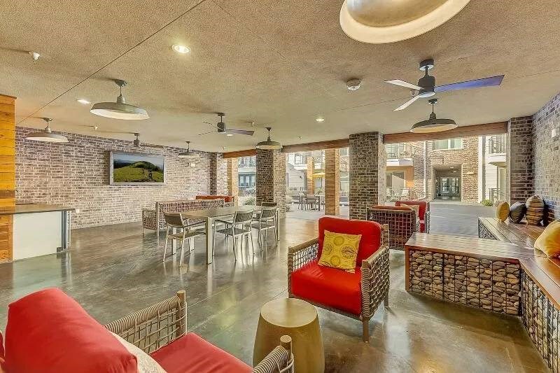 Spacious modern lounge here at Skyline at Farmers Market Apartment Homes with brick walls, featuring red cushioned chairs, a long table with gray chairs, ceiling fans, and bright natural light.