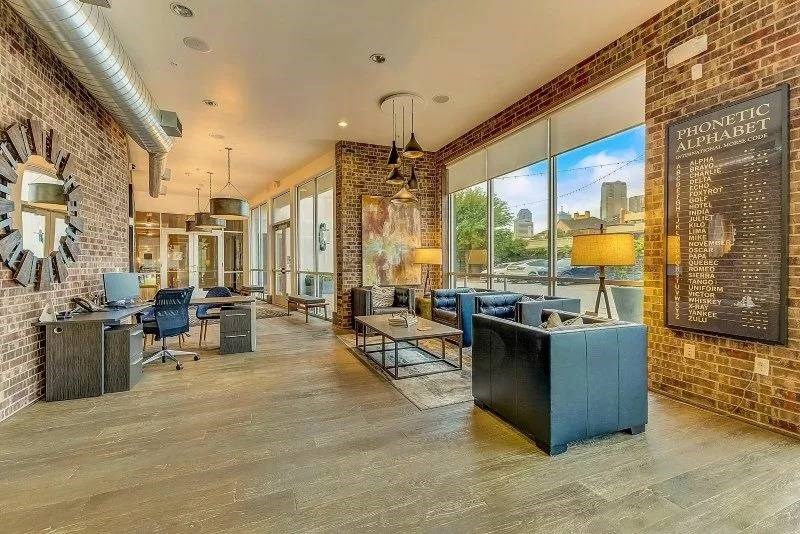 Trendy office here at Skyline at Farmers Market Apartment Homes with brick walls, large windows, industrial decor. Features workstations, lounge area, and a "Phonetic Alphabet" artwork. Warm, inviting ambiance.