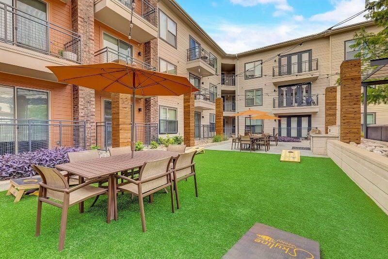 Courtyard here at Skyline at Farmers Market Apartment Homes with artificial grass, tables with orange umbrellas, beige chairs, and a cornhole game set. Surrounded by a three-story building.