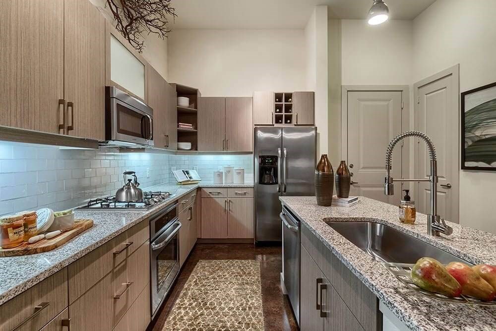 A modern kitchen here at Skyline at Farmers Market Apartment Homes with light wood cabinets, stainless steel appliances, and granite countertops. A centerpiece island holds pears and decor, under soft lighting.