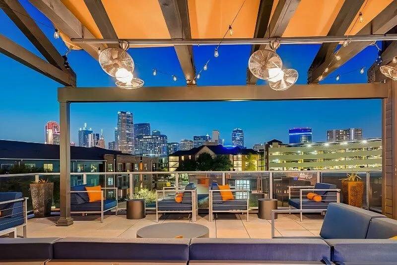Rooftop terrace here at Skyline at Farmers Market Apartment Homes with modern seating, orange cushions, string lights, and fans. Overlooks a city skyline at dusk with vibrant blue sky. Relaxed ambiance.