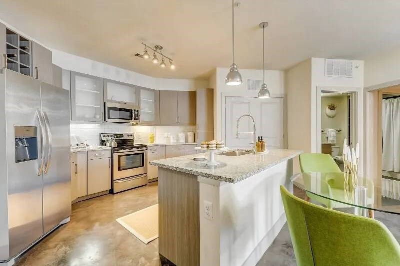 Modern kitchen here at Skyline at Farmers Market Apartment Homes with stainless steel appliances, a central island with granite countertop, two pendant lights, and a glass dining table with green chairs.