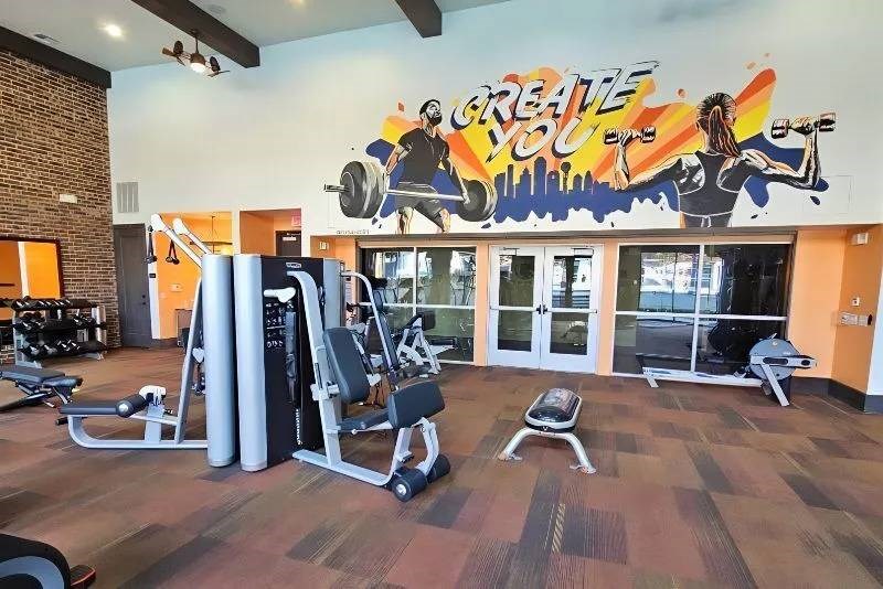 Modern gym interior here at Skyline at Farmers Market Apartment Homes with exercise equipment and a colorful mural reading "Create You." The mural features vibrant depictions of people lifting weights.