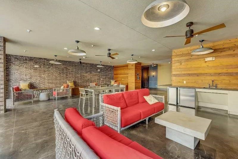 Modern lounge area here at Skyline at Farmers Market Apartment Homes with red cushioned sofas, brick walls, and wooden accents. Ceiling fans hang above, creating a cozy, contemporary vibe.