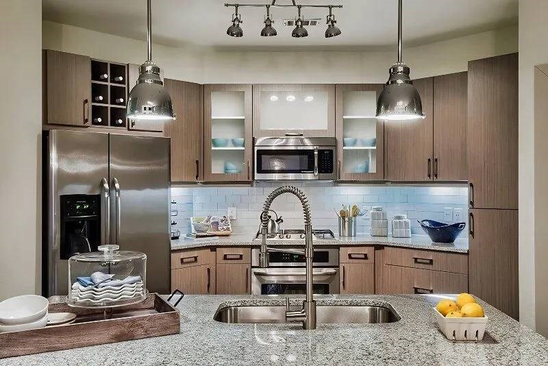 Modern kitchen here at Skyline at Farmers Market Apartment Homes with stainless steel appliances, granite countertops, and pendant lighting. Includes a double sink, lemons in a tray, and neatly arranged utensils.