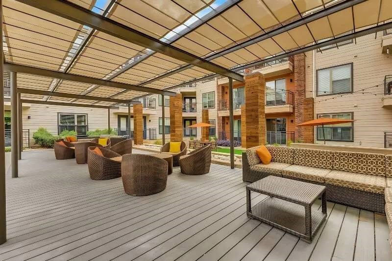 Spacious modern patio here at Skyline at Farmers Market Apartment Homes features wicker seating with orange and yellow cushions under a pergola, surrounded by apartment buildings and greenery, creating a cozy atmosphere.