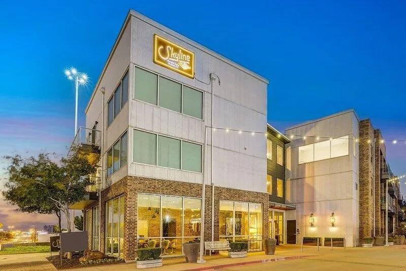 Modern building at dusk with a glowing "Skyline" sign. The structure features glass windows, warm lighting, and a welcoming ambiance against a vivid blue sky.