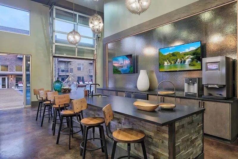 Modern lounge area here at Skyline at Farmers Market Apartment Homes with high ceilings and pendant lights. Features a bar counter with stools, wall-mounted TVs, a coffee machine, and decorative vases.