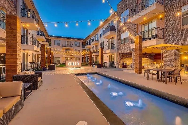 Courtyard here at Skyline at Farmers Market Apartment Homes at dusk with string lights, brick apartment buildings, cozy seating, and a fountain. A "Chill" sign adds a relaxed vibe.