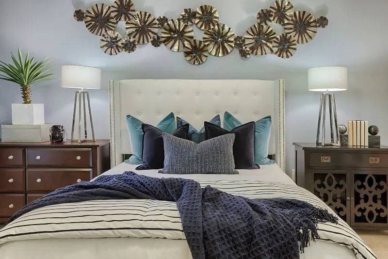 A cozy bedroom here at Skyline at Farmers Market Apartment Homes features a white, tufted headboard with blue and gray pillows. A navy throw is on the striped bedding. Modern lamps, a potted plant, and abstract wall art add elegance.