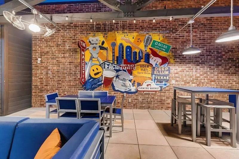 Outdoor patio here at Skyline at Farmers Market Apartment Homes with blue furniture. A colorful mural on the brick wall displays Dallas-themed images, exuding a lively, urban vibe. Hanging lights add a cozy touch.