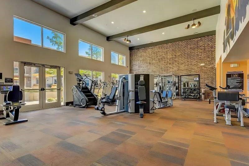Spacious gym here at Skyline at Farmers Market Apartment Homes with modern exercise equipment under high ceilings. Sunlight streams through large windows, illuminating treadmills and weight machines.