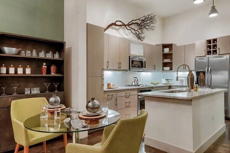 Modern kitchen here at Skyline at Farmers Market Apartment Homes with light wood cabinets and stainless steel appliances. A round glass table with green chairs sits in the foreground. Elegant and cozy ambiance.