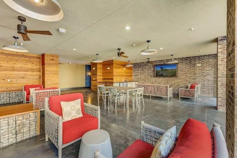 Spacious indoor lounge here at Skyline at Farmers Market Apartment Homes with modern decor; red-cushioned wicker sofas, wooden panels, brick walls, ceiling fans, creating a warm and welcoming atmosphere.