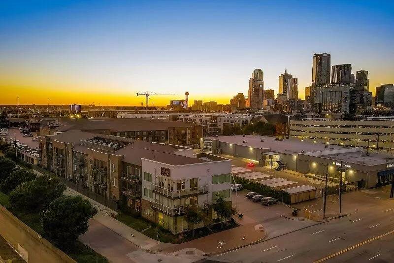 Sunset view of Skyline at Farmers Market Apartment Homes with illuminated buildings and construction cranes. In the foreground, modern low-rise buildings and parked cars line a quiet street.