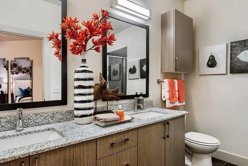 Modern bathroom here at Skyline at Farmers Market Apartment Homes with a granite countertop, dual sinks, and wall mirrors. A black and white striped vase with orange flowers adds a vibrant touch.