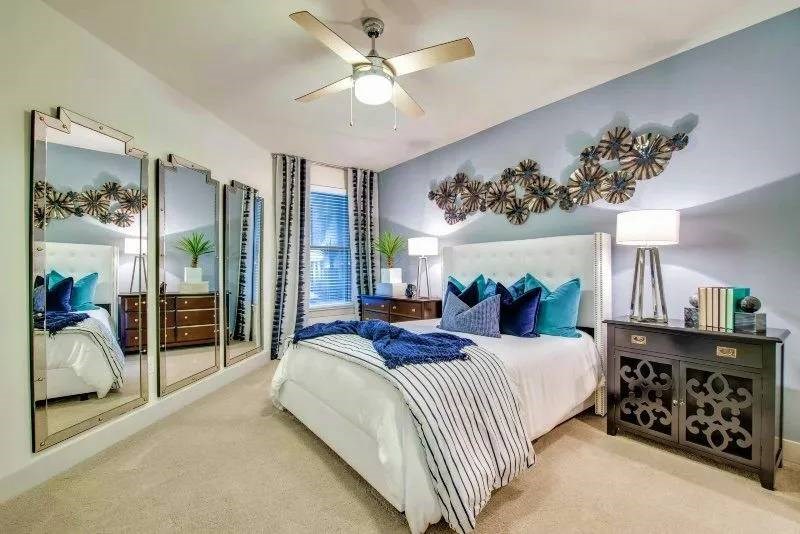 A stylish bedroom here at Skyline at Farmers Market Apartment Homes with a blue accent wall, adorned with abstract metal art. It features a plush bed with teal pillows, mirrored doors, and a ceiling fan.