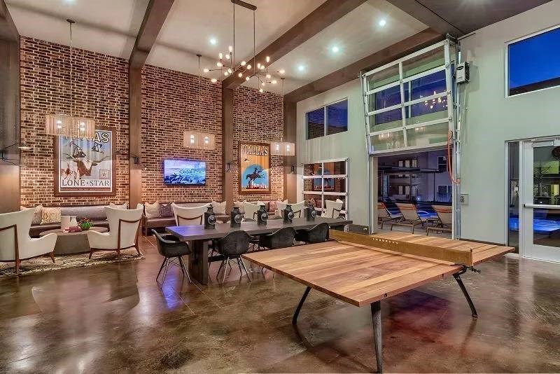 Spacious modern lounge here at Skyline at Farmers Market Apartment Homes with exposed brick walls, a long wooden dining table, chandeliers, and a ping pong table. Large windows reveal a night view.