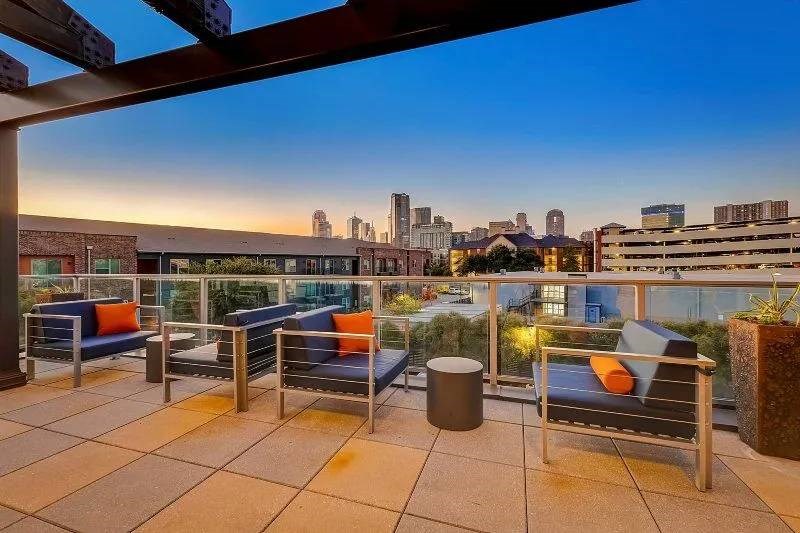 Rooftop patio here at Skyline at Farmers Market Apartment Homes at sunset featuring modern chairs with blue cushions and orange pillows. Overlooks a city skyline, creating a serene, urban ambiance.