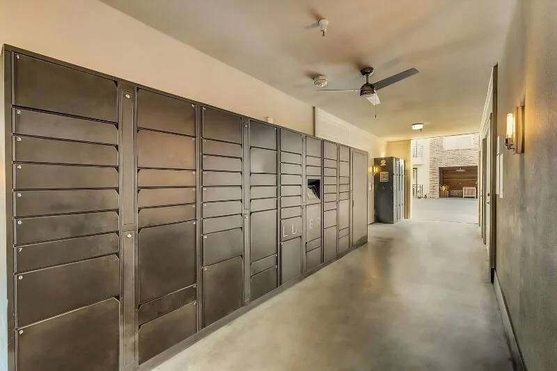 A row of large, black parcel lockers here at Skyline at Farmers Market Apartment Homes in a well-lit corridor, with a ceiling fan above. The space appears clean and modern, leading to an outdoor area.