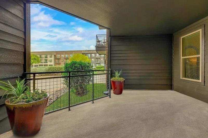 Covered patio here at Skyline at Farmers Market Apartment Homes with dark siding, potted plants in red pots, overlooking a grassy courtyard. Background shows an apartment building under a blue sky.