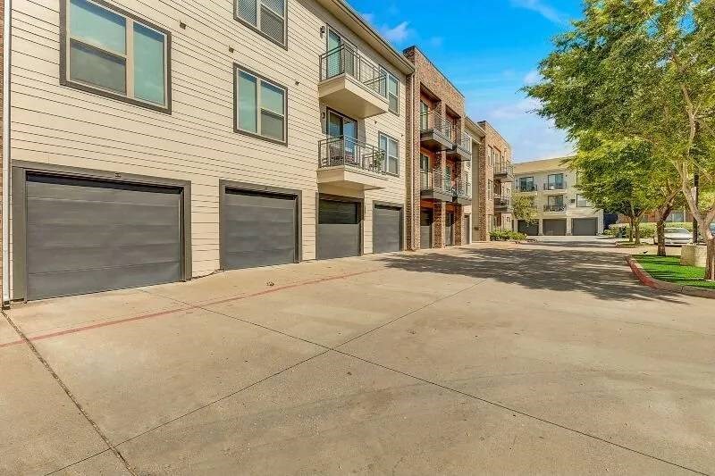 Modern Skyline at Farmers Market Apartment Homes with beige and brick facades, garages, and balconies, under a bright blue sky. A tree-lined driveway leads into the complex.