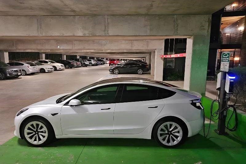 A white electric car is parked at a charging station in a dimly lit parking garage here at Skyline at Farmers Market Apartment Homes. Other vehicles are parked in rows, creating a calm, urban atmosphere.