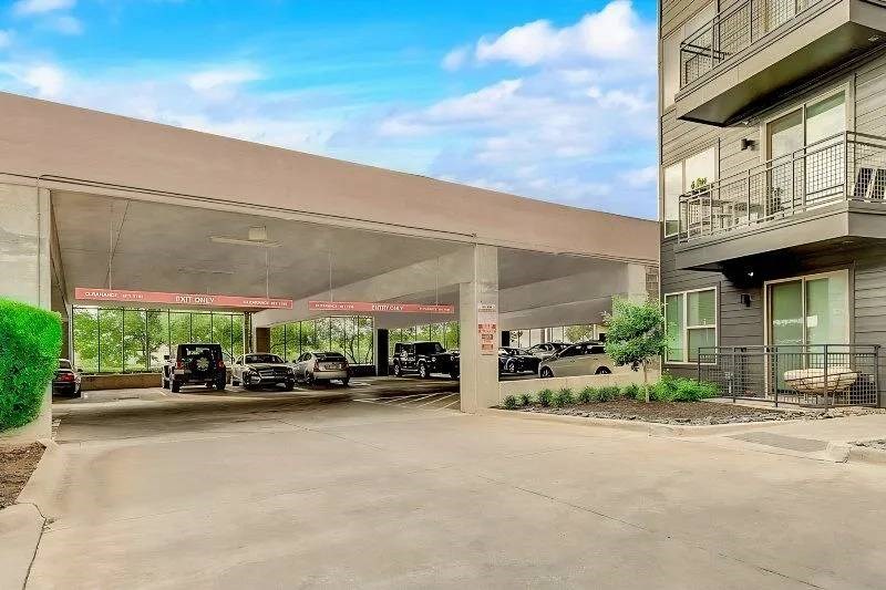Covered parking area here at Skyline at Farmers Market Apartment Homes with several cars parked beneath a concrete roof. Adjacent to an apartment building with balconies. Bright blue sky overhead.