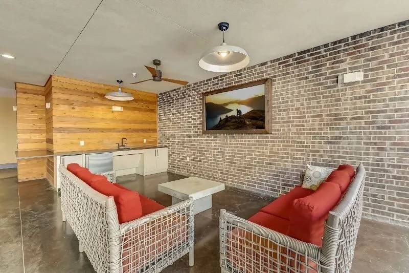Modern lounge here at Skyline at Farmers Market Apartment Homes with red-cushioned rattan sofas, a beige coffee table, and a brick wall featuring a landscape photo. Wood-panel accent and kitchenette in the back.