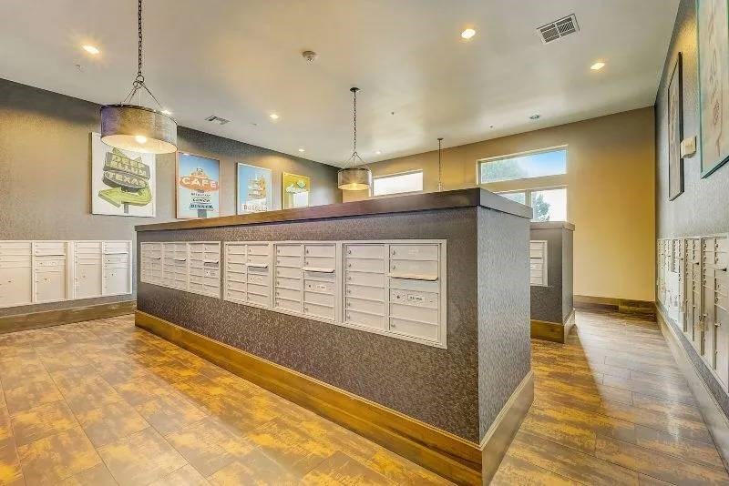 A modern mailroom here at Skyline at Farmers Market Apartment Homes with rows of white mail slots on textured gray walls. Warm lighting creates an inviting atmosphere, enhanced by wooden floors and framed art.