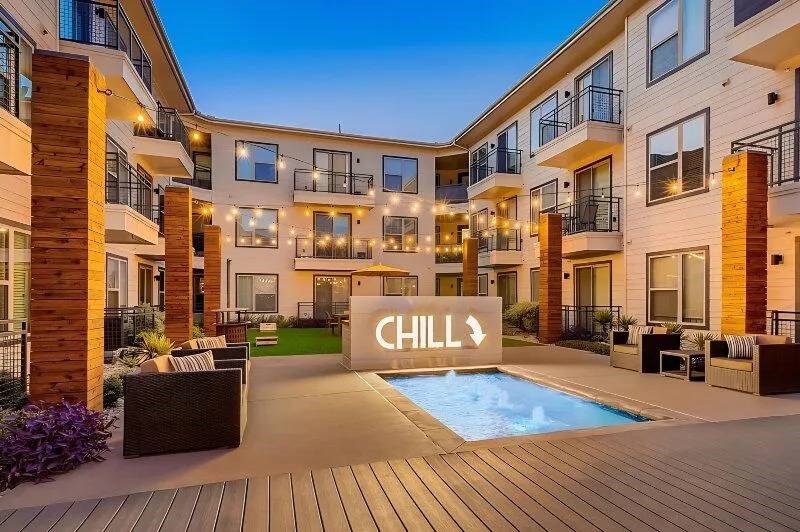 Modern courtyard here at Skyline at Farmers Market Apartment Homes with a small pool, cozy seating, and wooden columns. Illuminated sign reads "CHILL" at twilight, creating a relaxed atmosphere.