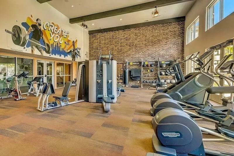 Spacious gym here at Skyline at Farmers Market Apartment Homes with cardio machines, weight equipment, and a mural saying "Create You." Sunlight streams through tall windows, creating an energetic vibe.