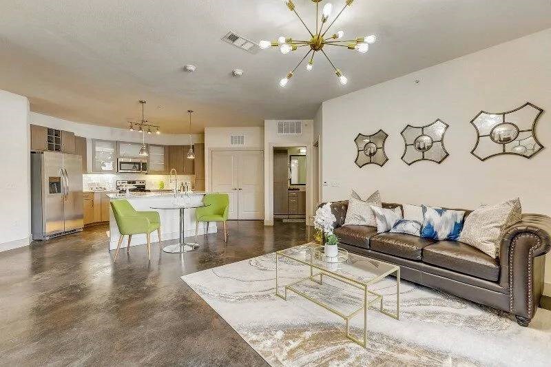 Modern living room here at Skyline at Farmers Market Apartment Homes with open kitchen. Features a brown leather sofa, glass coffee table, and green chairs. Bright lighting and abstract wall mirrors add elegance.
