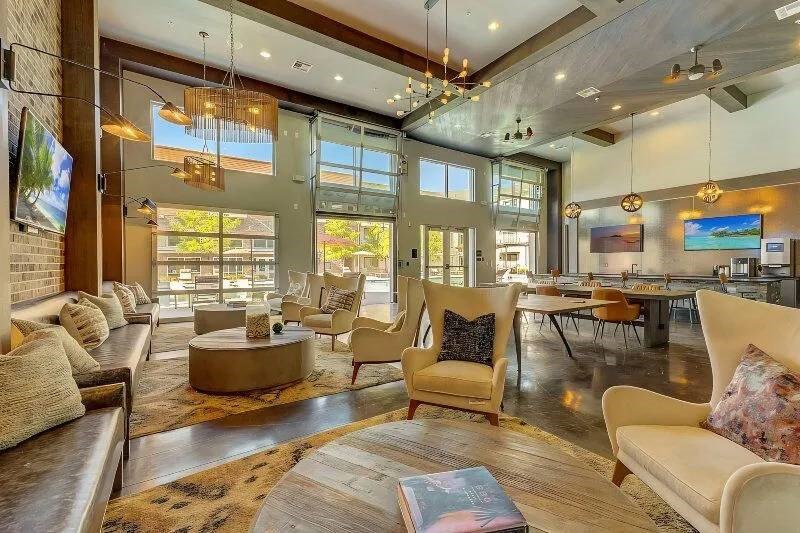 A spacious, modern lounge area here at Skyline at Farmers Market Apartment Homes with high ceilings, ample natural light, beige armchairs, wooden tables, and a cozy, relaxed ambiance.